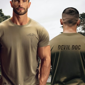 Puede incluir: Camisetas verde oliva con un símbolo médico y el texto "DEVIL DOC" en la espalda. Las camisetas están etiquetadas como "Military Green" en una fuente de escritura. Las camisetas son de manga corta y parecen estar hechas de un material suave.