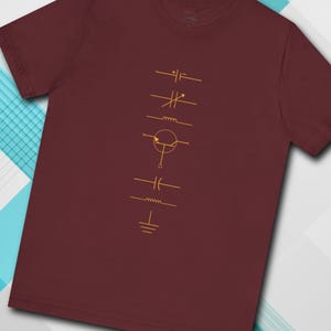 Può includere: T-shirt color bordeaux con una serie di simboli di circuiti elettronici dorati disposti verticalmente. I simboli includono un condensatore, un induttore e un resistore. La maglietta è di colore bordeaux unito.