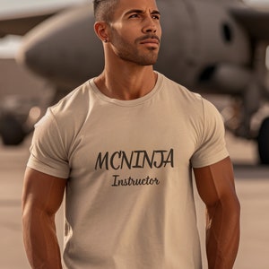 May include: A man wearing a beige t-shirt with the text "MCNINJA Instructor" printed on the front. He is standing in front of a large airplane.
