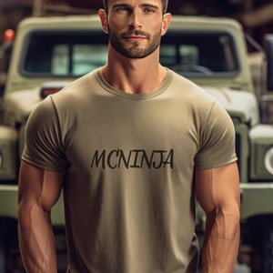 May include: A man wearing a khaki t-shirt with the text "MCNINJA" printed on the front. He is standing in front of a green military vehicle.