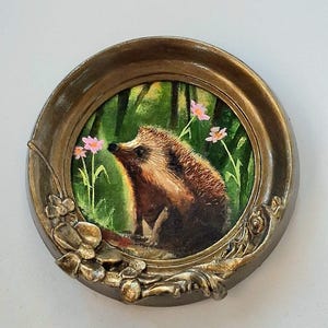 Puede incluir: Un marco circular ornamentado de color bronce con detalles florales en relieve en la parte inferior. Dentro del marco hay una pintura de un erizo marr&oacute;n en un entorno de bosque verde con peque&ntilde;as flores rosas visibles al fondo.