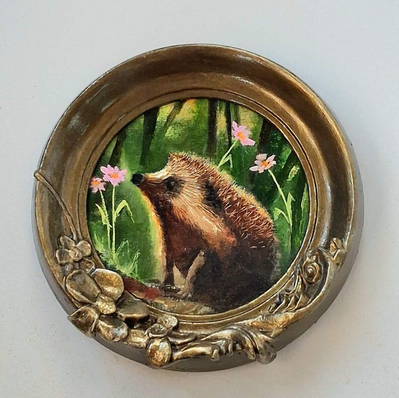 Puede incluir: Un marco circular ornamentado de color bronce con detalles florales en relieve en la parte inferior. Dentro del marco hay una pintura de un erizo marr&oacute;n en un entorno de bosque verde con peque&ntilde;as flores rosas visibles al fondo.