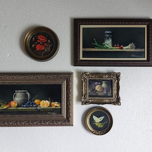 K&ouml;nnte beinhalten: Eine Sammlung gerahmter Stillleben-Gem&auml;lde. Die Kunstwerke zeigen verschiedene Motive, darunter Blumen, Fr&uuml;chte und Gem&uuml;se. Die Rahmen sind verziert und in Braun- und Goldt&ouml;nen gehalten. Die Gem&auml;lde h&auml;ngen an einer wei&szlig;en Wand.