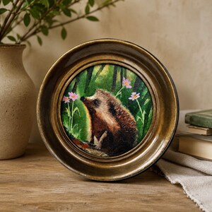 Puede incluir: Una pintura circular de un erizo marr&oacute;n en un entorno de bosque verde con peque&ntilde;as flores rosas, encerrada en un marco redondo de metal color oro antiguo con un acabado liso y pulido y un borde interior oscuro, presentada como arte mural.