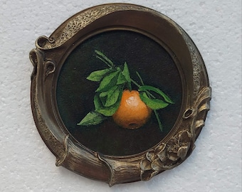 Puede incluir: Una pequeña pintura redonda de una fruta naranja con hojas verdes, sobre un fondo oscuro. La obra de arte está enmarcada en un marco decorativo de bronce de estilo antiguo, lo que añade una estética vintage. La pintura captura los colores y texturas naturales de la fruta.