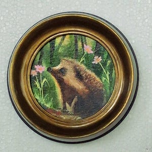 Puede incluir: Una pieza de arte mural circular que presenta una pintura de un erizo marr&oacute;n en un entorno de bosque verde con peque&ntilde;as flores rosas. La obra est&aacute; encerrada en un marco de metal dorado pulido con un perfil de borde redondeado.