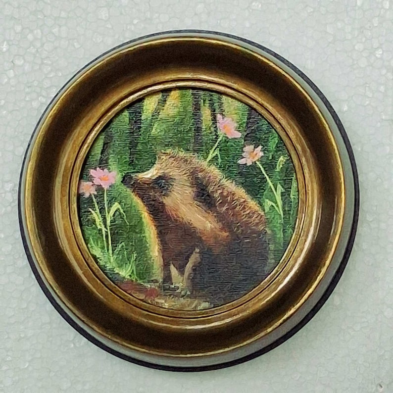 Puede incluir: Una pieza de arte mural circular que presenta una pintura de un erizo marr&oacute;n en un entorno de bosque verde con peque&ntilde;as flores rosas. La obra est&aacute; encerrada en un marco de metal dorado pulido con un perfil de borde redondeado.