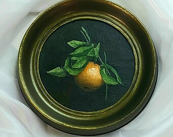 Puede incluir: Una pintura circular de una naranja con hojas verdes centrada sobre un fondo oscuro, encerrada en un marco redondo de metal dorado con acabado texturizado. La obra de arte presenta sombreados realistas y un contraste de colores vivo.