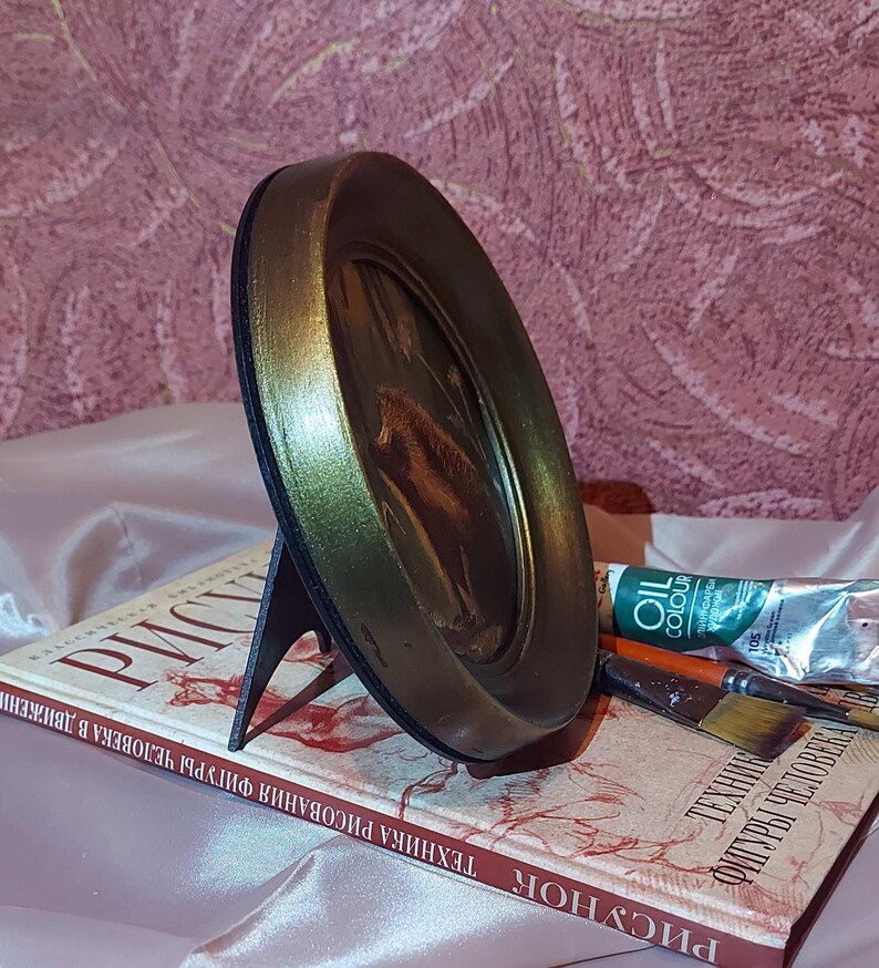 Puede incluir: Un marco de fotos redondo de color dorado met&aacute;lico con soporte negro, que contiene una pintura oscura de un caballo. El marco descansa sobre un libro con t&iacute;tulo en cir&iacute;lico, junto a un tubo de &oacute;leo verde y un pincel de mango de madera.