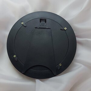 Puede incluir: Una parte trasera de marco de fotos circular de color negro que cuenta con un soporte de caballete central articulado para exhibici&oacute;n en mesa y un colgador de metal para montaje en pared. Cuatro clips giratorios negros aseguran el panel.