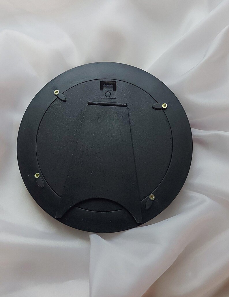 Puede incluir: Una parte trasera de marco de fotos circular de color negro que cuenta con un soporte de caballete central articulado para exhibici&oacute;n en mesa y un colgador de metal para montaje en pared. Cuatro clips giratorios negros aseguran el panel.