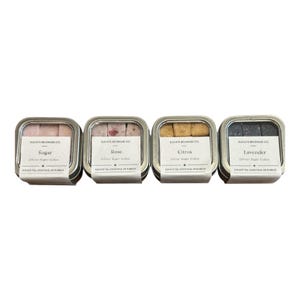 May include: Four small, square metal tins, each containing sugar cubes. The tins are labeled "Sugar", "Rose", "Citrus", and "Lavender". The text "Elevate Beverage Co." and "Clover Sugar Cubes" are also visible on the labels.