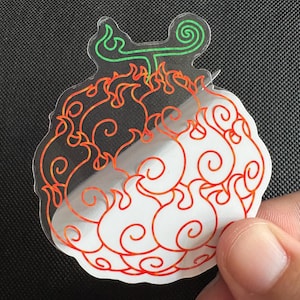 May include: A clear sticker featuring a stylized fruit design with orange and red flame-like patterns. The top of the fruit has green accents. The sticker is held by a hand against a dark background.