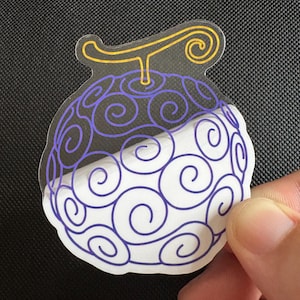 May include: A clear sticker featuring a stylized fruit design with purple swirls and a yellow stem. The sticker has a white base and is held against a dark background.
