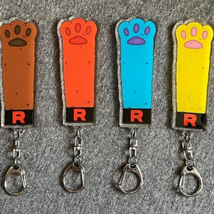 Puede incluir: Cuatro llaveros con huellas de patas de diferentes colores. Las huellas de patas son marrones, rojas, azules y amarillas. Cada llavero tiene una etiqueta negra con la letra "R" en ella.