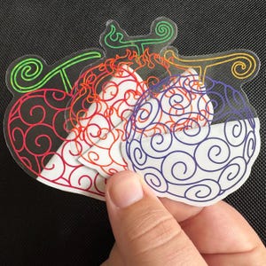 May include: A hand holds a stack of transparent stickers featuring colorful, stylized fruit designs. Each sticker has a different color outline and internal swirl pattern, including red, orange, green, and purple. The stickers are on a black background.