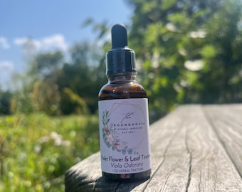 Puede incluir: Una botella de vidrio marrón con un gotero, etiquetada con "The Roundhouse Herbal Medicine Est. 2023" y "Violet Flower & Leaf Tincture Viola Odorata 1 oz Herbal Tincture". La botella está sobre una superficie de madera con un fondo borroso de follaje verde y un cielo azul.