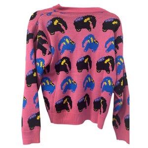 May include: Pink knit sweater featuring a repeating pattern of pixelated elephants in black, blue, and yellow. The sweater has a crew neck and long sleeves. The elephants are arranged in a grid-like pattern across the fabric.
