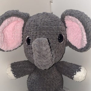 May include: A gray crocheted elephant plush toy with pink ears and white feet. The elephant has black eyes and a gray trunk.