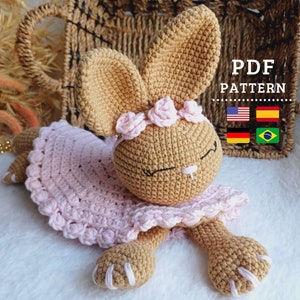 May include: A crocheted bunny wearing a pink dress with a flower crown. The bunny is lying on a white surface. The image includes the text "PDF PATTERN" and flags for the United States, Spain, Germany, and Brazil.