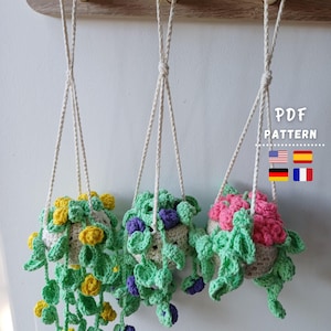 May include: Three handmade crochet hanging planters with trailing vines and flowers in yellow, purple, and pink. The planters are suspended by white cords from a wooden shelf. The text "PDF PATTERN" is visible in the image.