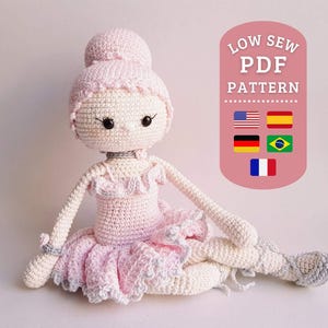 Puede incluir: Una muñeca bailarina de crochet con un vestido rosa y un moño. La muñeca tiene ojos negros y una gargantilla negra. La muñeca está sentada sobre una superficie blanca. La imagen incluye el texto "LOW SEW PDF PATTERN" y banderas de Estados Unidos, España, Alemania, Brasil y Francia.