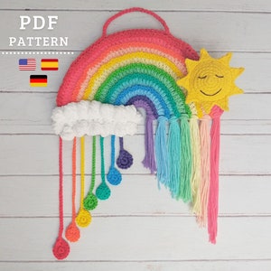 Może przedstawiać: Wiszący dekoracyjny tęcza wykonany na szydełku z uśmiechniętym słońcem i puszystą chmurką z kroplami deszczu. Tęcza wykonana jest z kolorowej włóczki i ma frędzle zwisające z dołu. Słońce jest żółte i ma uśmiechniętą buźkę z zamkniętymi oczami. Chmurka jest biała i ma kilka kropel deszczu zwisających z niej.