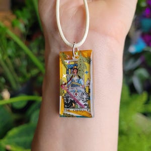 May include: A rectangular pendant necklace featuring a tarot card design. The pendant is yellow with a tarot card image of a person in a chariot. The pendant is adorned with dried flowers and glitter, suspended on a cream-colored cord.