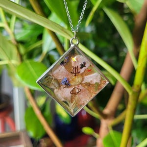 May include: A silver chain necklace with a square pendant. The pendant is clear with a floral design and small embellishments, including a star and other symbols. The background is blurred green foliage.