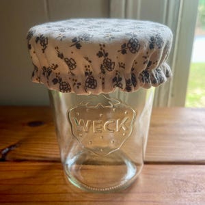 May include: A clear glass jar with the embossed word "WECK" on the front. The jar is topped with a fabric lid featuring a white background and a pattern of small, dark floral designs. The jar sits on a wooden surface.