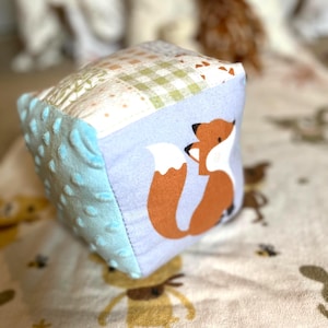 May include: A soft, multi-textured baby block featuring a fox design. The cube has light blue, patterned, and fox-printed sides. The fox is orange with a white tail. The block is designed for babies and toddlers.