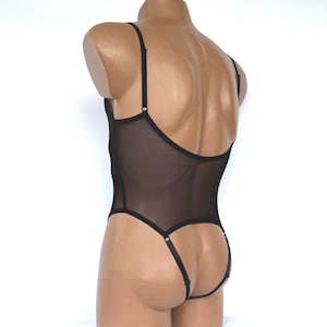 Puede incluir: Body negro transparente con tirantes ajustables y espalda de tanga. La lencería presenta un escote redondo y un tejido de malla delicado y transparente. La prenda se exhibe en un maniquí sobre un fondo blanco.