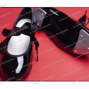 May include: A pair of black leather tap shoes with a black ribbon tied around the ankle of one shoe. The shoes are on a red background.