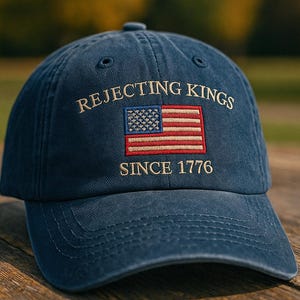Può includere: Un cappellino da baseball blu navy con la scritta ricamata "REJECTING KINGS SINCE 1776" e una toppa con la bandiera americana. Il cappello è realizzato in un tessuto morbido e poggia su una superficie di legno.