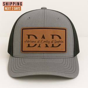 May include: Gray trucker hat with a black mesh back and a brown leather patch. The patch features the word "DAD" and the names "Adriana & Emily & Sophia" in a stylish font.