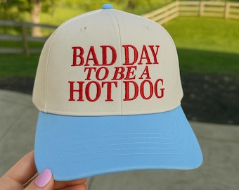 May include: A baseball cap with a cream-colored crown and a light blue brim. The cap features the embroidered phrase "BAD DAY TO BE A HOT DOG" in red lettering. The cap has a rounded top and a curved brim.