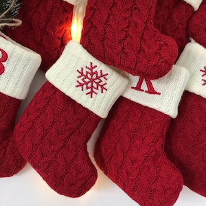 Peut inclure: Gros plan sur plusieurs bas de Noël rouges tricotés avec des revers blancs. Les bas ont un motif en tricot torsadé et sont ornés de flocons de neige et de lettres brodés. Les bas sont éclairés par des lumières blanches chaudes, créant une scène de vacances festive.