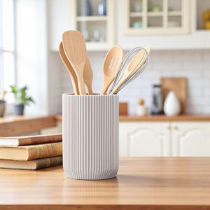 以下が含まれることがあります： 木製のカウンターの上に置かれた、薄いグレーのリブ付きの調理器具ホルダー。木のスプーンと泡立て器が入っています。調理器具は、窓と本の山がある明るい白いキッチンにあります。