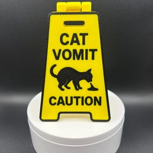 May include: A yellow and black caution sign with the words "CAT VOMIT" and a silhouette of a cat vomiting. The sign is in the shape of a traditional caution sign with a folding hinge at the top.
