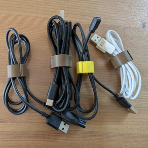 Puede incluir: Varios cables de carga USB, incluyendo cables negros y blancos, sujetos con bridas marrones y amarillas. Los cables están dispuestos sobre una superficie de madera marrón claro.
