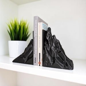 May include: Black mountain-shaped bookends with a textured surface, holding two books on a white shelf. The bookends have a mountain range design. A potted plant is in the background.