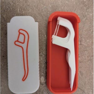 May include: A white dental floss pick case with an orange outline of a floss pick on the front. The case is open, revealing a stack of white floss picks inside an orange container. The case and picks are on a gray surface.