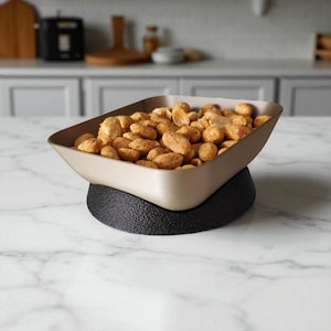 May include: A beige serving bowl filled with roasted nuts sits atop a textured black base. The bowl has a rectangular shape with slightly angled sides. The nuts are golden brown and appear to be seasoned.