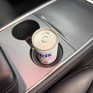 Puede incluir: Una lata de bebida con sabor a fresa "Ikis" está en el portavasos de un coche. La lata blanca tiene gráficos rosas y azules. El interior del coche es negro y gris, con molduras plateadas y un lápiz óptico integrado.