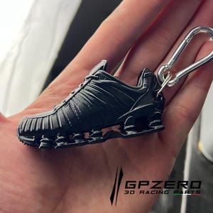 Può includere: Un portachiavi a forma di sneaker nera in miniatura con un moschettone argentato. La scarpa presenta un design dettagliato, inclusa la suola e i lacci. Il testo "GPZERO 3D RACING PARTS" è visibile in basso.