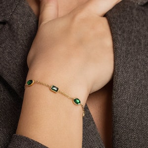 以下が含まれることがあります： 3つの緑色の宝石のアクセントが付いた金のチェーンブレスレット。ブレスレットは繊細なチェーンと楕円形と長方形の石を持っています。