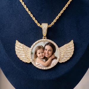 May include: A gold-toned circular pendant necklace featuring a central photograph of a child and an adult. The pendant is framed by sparkling stones and accented with two symmetrical, stone-encrusted wings on either side, suspended from a rope chain.