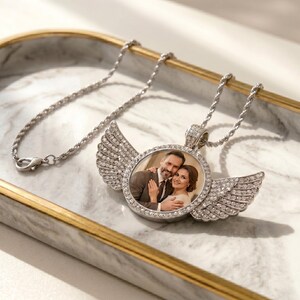 May include: A silver-toned circular pendant necklace featuring a central photograph of a couple. The pendant is framed by clear crystals and adorned with two symmetrical, crystal-encrusted angel wings on either side, attached to a twisted chain.