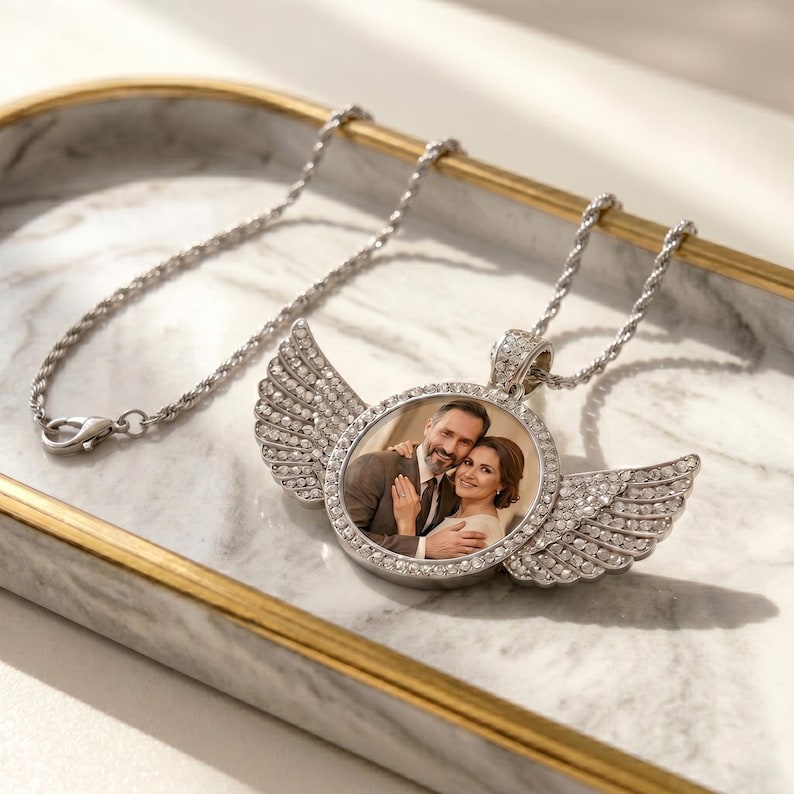 May include: A silver-toned circular pendant necklace featuring a central photograph of a couple. The pendant is framed by clear crystals and adorned with two symmetrical, crystal-encrusted angel wings on either side, attached to a twisted chain.