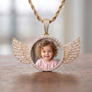 May include: A gold-toned circular pendant necklace featuring a central photograph of a curly-haired child wearing a pink shirt. The pendant is framed by clear stones and adorned with two symmetrical, stone-encrusted angel wings on each side.